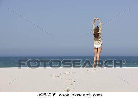Stock Photo - Rear view of a woman standing on the beach with her arms raised. Fotosearch