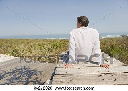 Rear view of a man sitting on a boardwalk on the beach View Large Photo Image Stock Image - Rear view of a man sitting on a boardwalk on the beach. Fotosearch