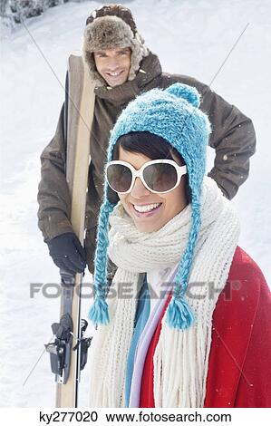 Stock Image - Young woman in winter clothes smiling at camera, man holding skis in background. Fotosearch