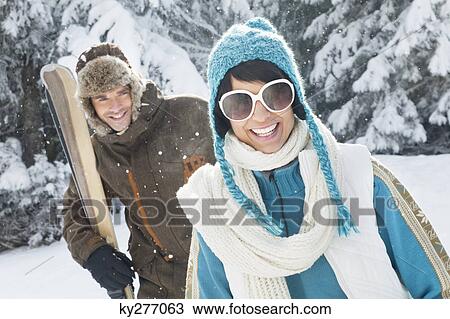 Stock Image - Young woman in winter clothes smiling at camera, man holding skis in background. Fotosearch