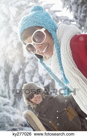 Young woman in winter clothes smiling at camera, man holding skis in background View Large Photo Image Stock Photograph - Young woman in winter clothes smiling at camera, man holding skis in background. Fotosearch