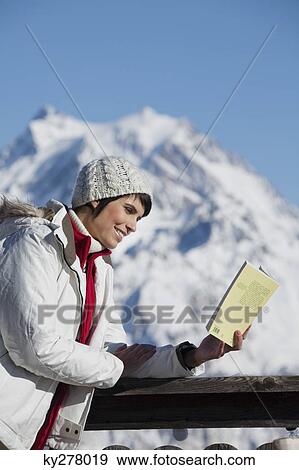 Stock Photo - Young woman reading a book, enjoying winter sun, mountains in background. Fotosearch