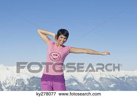 Stock Photo - Young woman with arms raised, smiling at camera, mountains in background. Fotosearch