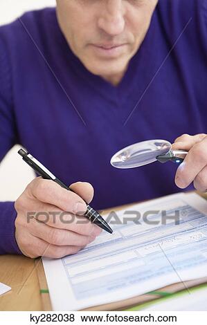 Stock Photo - Man looking through a magnifying glass while filling his tax form. Fotosearch