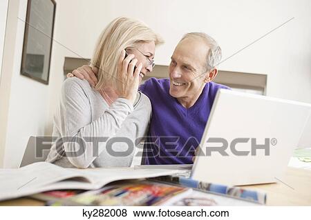 Stock Photo - Woman talking on a mobile phone sitting with her husband in front of a laptop. Fotosearch
