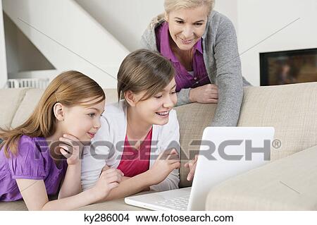 Two girls using a laptop on a couch with their grandmother standing beside them View Large Photo Image Picture - Two girls using a laptop on a couch with their grandmother standing beside them. Fotosearch