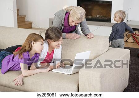 Stock Image - Two girls using a laptop on a couch with their grandmother standing beside them. Fotosearch