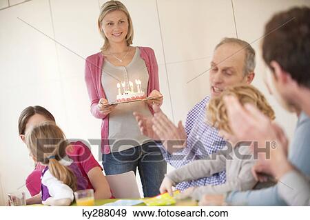 Stock Photo - Family at a birthday celebration with a woman bringing cake in the background. Fotosearch