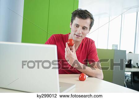 Stock Image - Businessman holding a tomato and using a laptop. Fotosearch