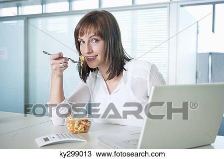 Stock Image - Businesswoman eating pasta in front of a laptop. Fotosearch