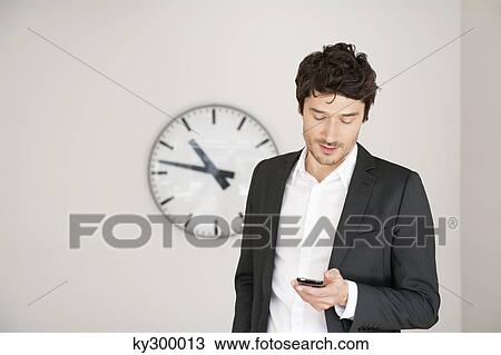 Businessman standing in front of a clock and using a mobile phone View Large Photo Image Stock Image - Businessman standing in front of a clock and using a mobile phone. Fotosearch