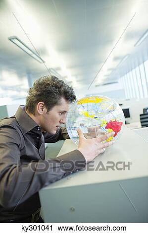 Stock Image - Businessman holding a globe and thinking in an office. Fotosearch