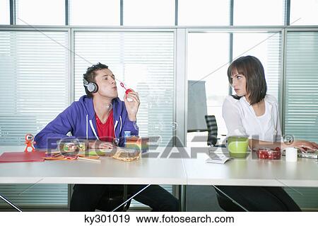 Businesswoman looking at a businessman listening to headphones and blowing bubbles View Large Photo Image Stock Photo - Businesswoman looking at a businessman listening to headphones and blowing bubbles. Fotosearch