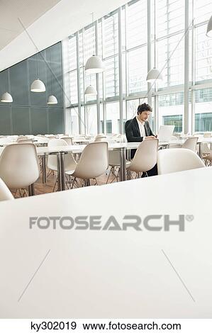Businessman sitting at a cafeteria and using a laptop View Large Photo Image Stock Photo - Businessman sitting at a cafeteria and using a laptop. Fotosearch