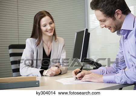Man signing documents with a female real estate agent sitting in front of him View Large Photo Image Picture - Man signing documents with a female real estate agent sitting in front of him. Fotosearch
