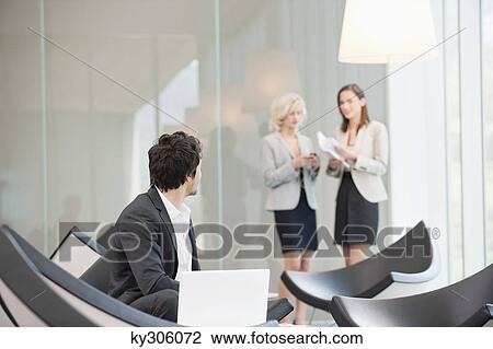 Stock Image - Businessman sitting in a waiting room looking at his colleagues standing in the background. Fotosearch