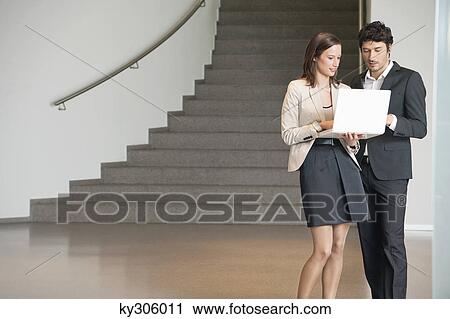 Stock Image - Two business executives standing in an office and using a laptop. Fotosearch