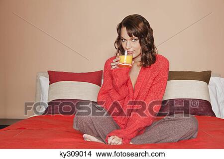 Picture - Young woman drinking orange juice, sitting cross-legged in bed. Fotosearch