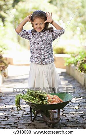 Stock Photo - Cute girl standing near a wheelbarrow filled with carrots. Fotosearch