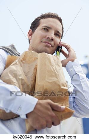 Stock Photography - Mid adult man talking on a mobile phone with paper bags full of vegetables. Fotosearch