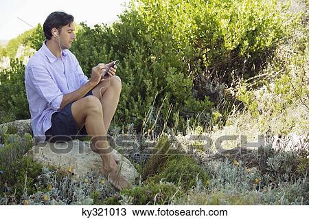 Mid adult man sitting on a rock and listening to music with an MP3 player View Large Photo Image Stock Image - Mid adult man sitting on a rock and listening to music with an MP3 player. Fotosearch