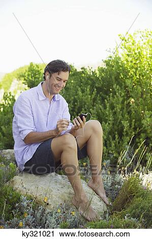 Mid adult man sitting on a rock and listening to music with an MP3 player View Large Photo Image Stock Image - Mid adult man sitting on a rock and listening to music with an MP3 player. Fotosearch