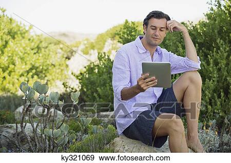 Mid adult man sitting on a rock and using a digital tablet View Large Photo Image Stock Photo - Mid adult man sitting on a rock and using a digital tablet. Fotosearch