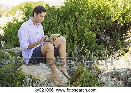 Mid adult man sitting on a rock and using a digital tablet View Large Photo Image Stock Photograph - Mid adult man sitting on a rock and using a digital tablet. Fotosearch