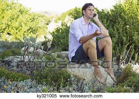 Mid adult man sitting on a rock and talking on a mobile phone View Large Photo Image Stock Photography - Mid adult man sitting on a rock and talking on a mobile phone. Fotosearch