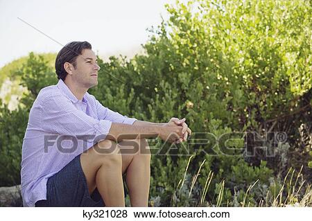 Stock Photo - Mid adult man sitting on a rock in a garden. Fotosearch