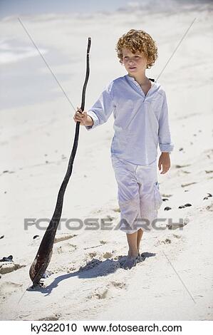Stock Image - Boy standing with a wooden stick on the beach. Fotosearch
