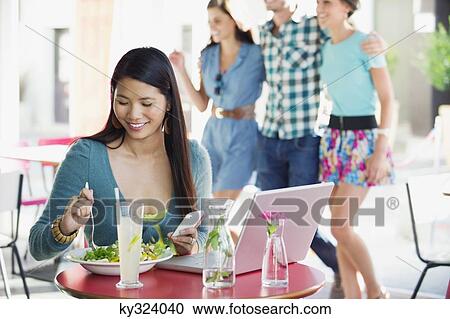 Smiling woman eating food while using mobile with people in the background at a restaurant View Large Photo Image Stock Image - Smiling woman eating food while using mobile with people in the background at a restaurant. Fotosearch