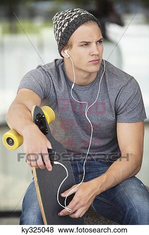 Man holding a skateboard and listening to music on a mobile phone View Large Photo Image Stock Photo - Man holding a skateboard and listening to music on a mobile phone. Fotosearch