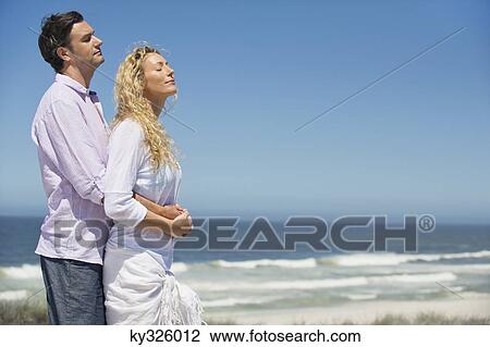Stock Image - Man embracing a woman from behind on the beach. Fotosearch