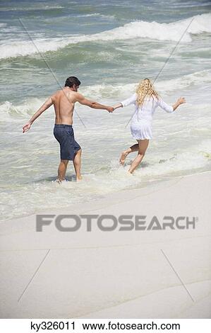 Stock Image - Rear view of a couple running on the beach. Fotosearch