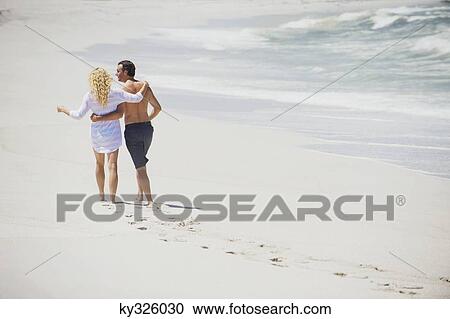 Stock Image - Rear view of a couple walking on the beach with their arms around each other. Fotosearch