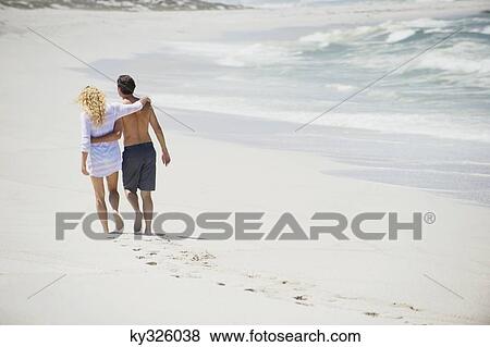 Stock Photo - Rear view of a couple walking on the beach with their arms around each other. Fotosearch