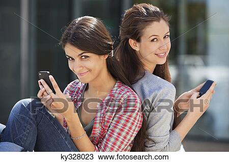 Portrait of two young women sitting back to back and using mobile phones View Large Photo Image Stock Image - Portrait of two young women sitting back to back and using mobile phones. Fotosearch