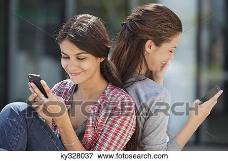 Two young women sitting back to back and text messaging View Large Photo Image Stock Photo - Two young women sitting back to back and text messaging. Fotosearch