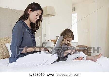 Stock Image - Woman working on a laptop with her daughter playing with a mobile phone beside her. Fotosearch