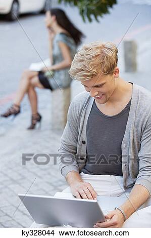 Picture - Young man using a laptop with a woman in the background on a street. Fotosearch