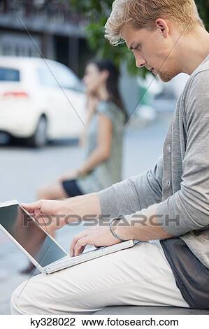 Young man using a laptop with a woman in the background on a street View Large Photo Image Stock Image - Young man using a laptop with a woman in the background on a street. Fotosearch