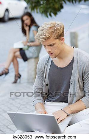 Young man using a laptop with a woman in the background on a street View Large Photo Image Stock Image - Young man using a laptop with a woman in the background on a street. Fotosearch
