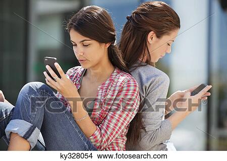 Young woman sitting back to back and text messaging View Large Photo Image Picture - Young woman sitting back to back and text messaging. Fotosearch