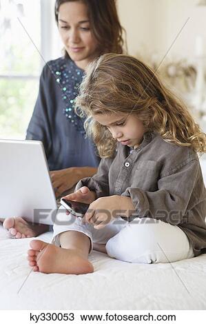 Girl looking at a smart phone with her mother working on a laptop on the bed View Large Photo Image Stock Image - Girl looking at a smart phone with her mother working on a laptop on the bed. Fotosearch