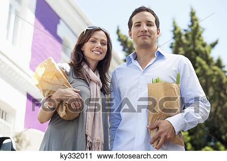 Stock Image - Portrait of a couple standing with paper bags full of vegetables. Fotosearch