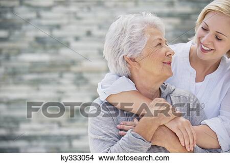 Picture - Young woman embracing her mother from behind and smiling. Fotosearch
