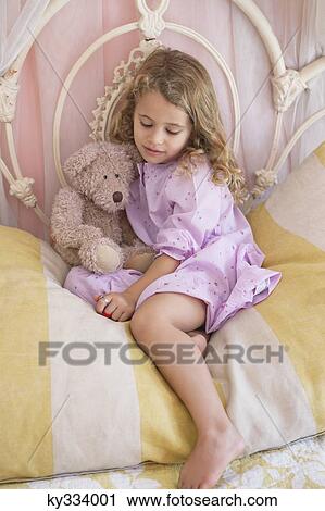 Stock Image - Cute little girl sitting with teddy bear on the bed. Fotosearch