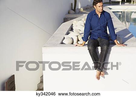 Stock Photograph - Mid adult man sitting on the terrace near a swimming pool. Fotosearch