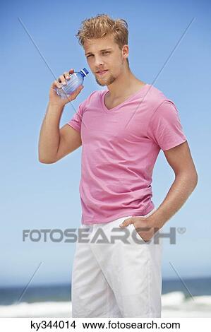 Picture - Portrait of a young man drinking water from a water bottle on the beach. Fotosearch